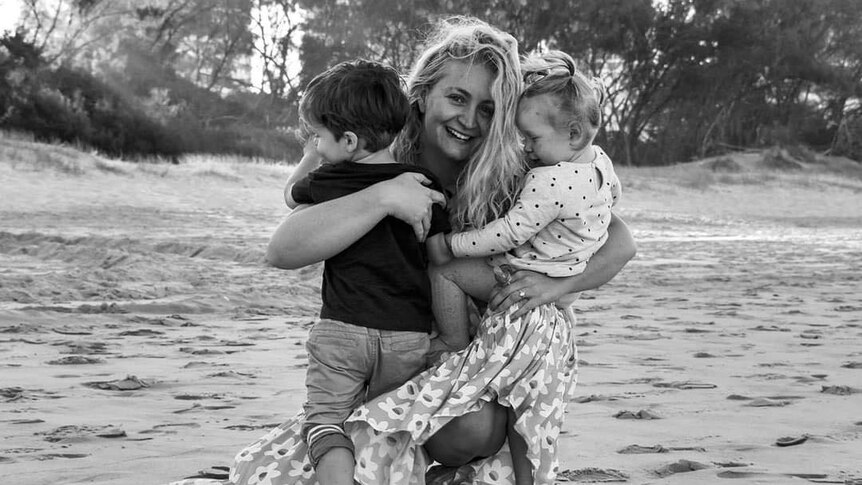 A black and white photo of a woman crouched down on a sand beach embracing two young children in her arms. 