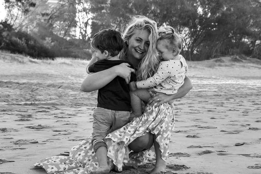 A black and white photo of a woman crouched down on a sand beach embracing two young children in her arms. 