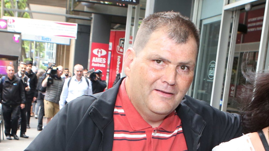 Michael Rogers, wearing a black jacket over a red polo shirt with white stripes, walks down a CBD street.