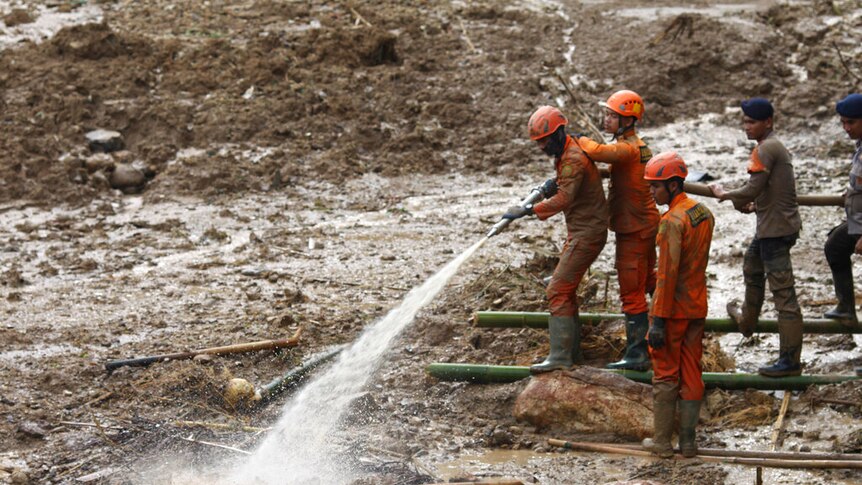 You view a muddy landscape as rescue workers in orange hold a hose which is spraying a clear liquid onto debris.