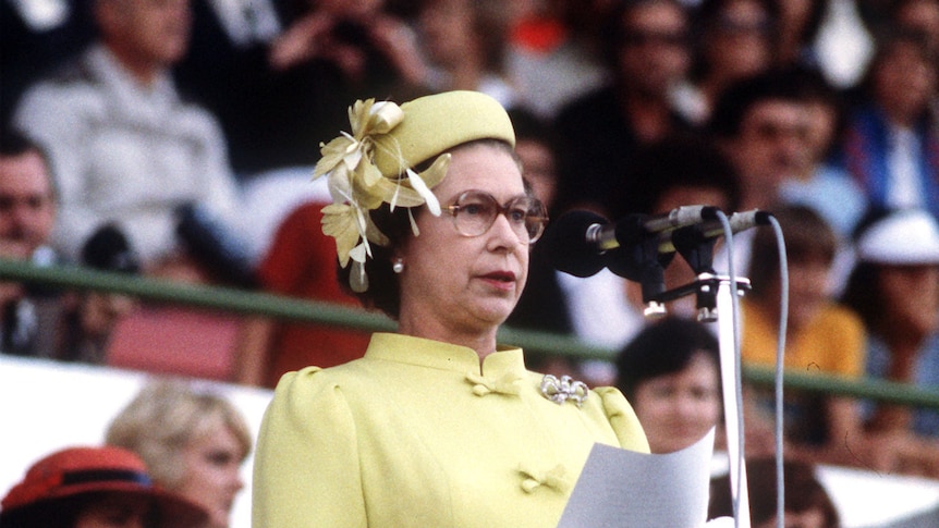 Queen Elizabeth stands and reads a speech into a microphone, crowds in a grandstand behind her.