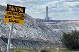 A sign saying "caution" in front of an open pit coal mine and power station.