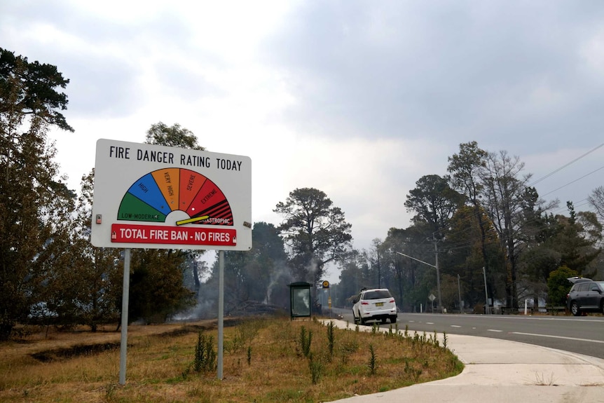 A fire danger rating sign by the roadside is pointed to 'catastrophic' as smoking embers can be seen behind.