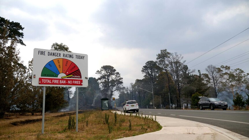 A fire danger rating sign by the roadside is pointed to 'catastrophic' as smoking embers can be seen behind.