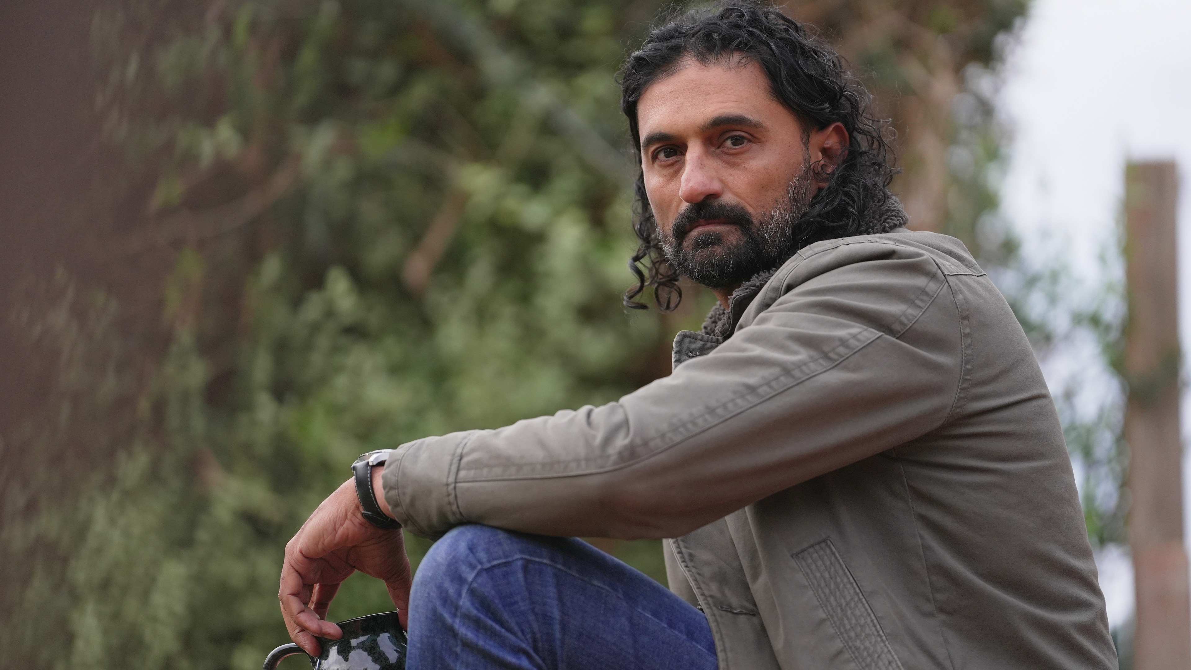 Man with shoulder-length hair and with facial hair sits turned slightly sideways. He is outdoors and wears a jacket and jeans.