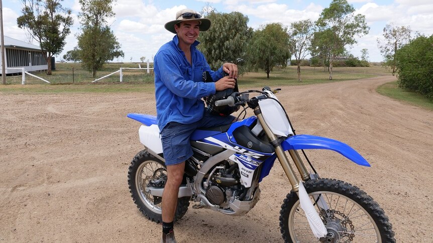 Tom Boyle sits on a blue and white motorbike holding a black helmet.
