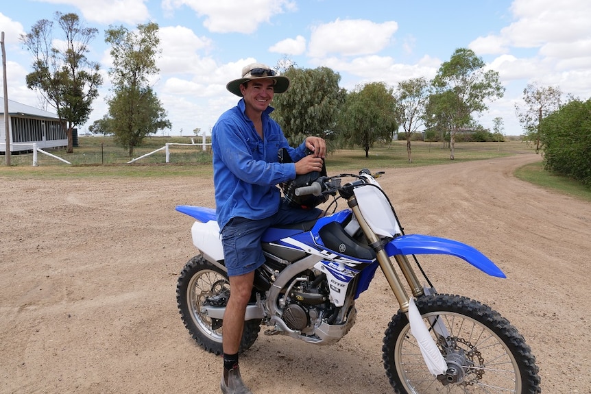 Tom Boyle sits on a blue and white motorbike holding a black helmet.