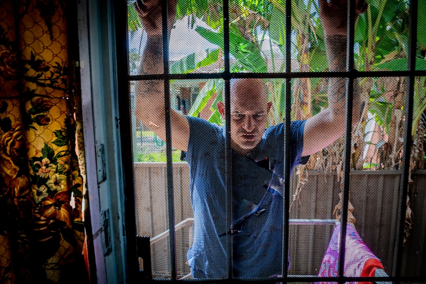 Man behind a flyscreen door with his arms up against it.