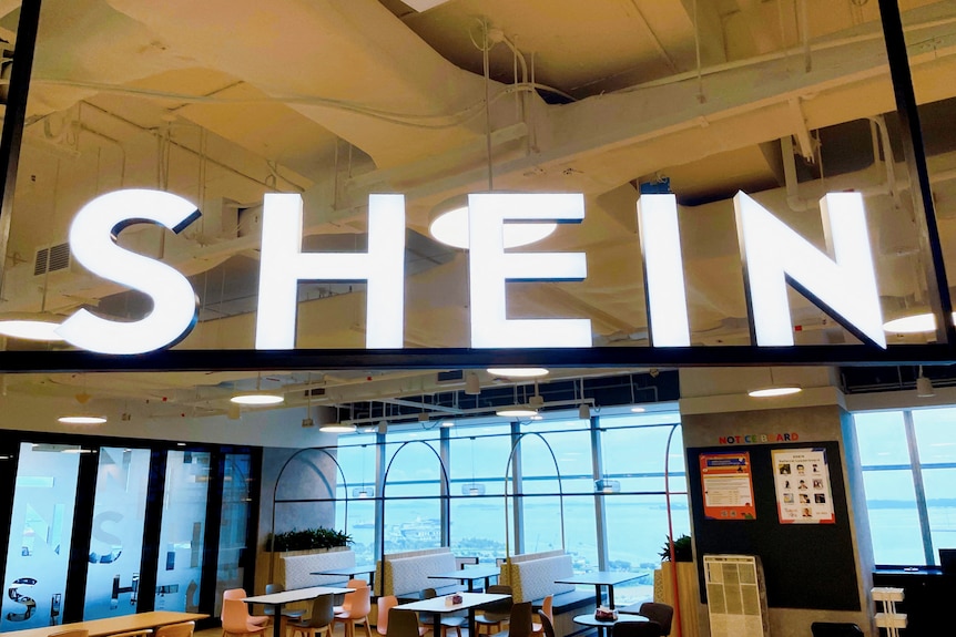 A fluorescent white logo that says Shein suspended from the roof of a commercial office.