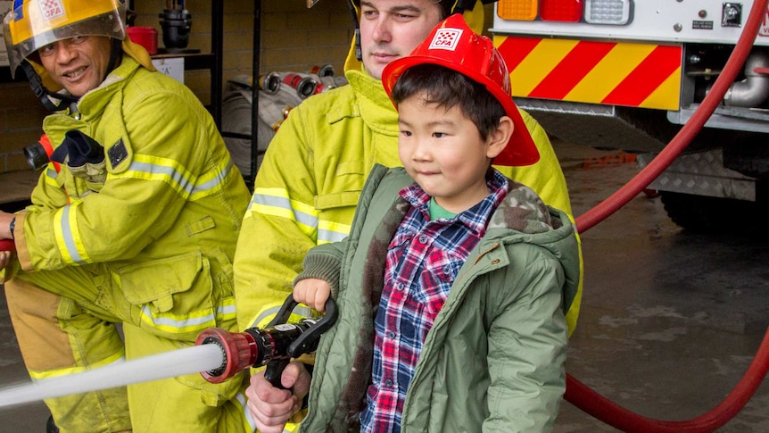 A young boy wearing a fire helmet uses a hose attached to a fire truck with the help of uniformed fire fighters.