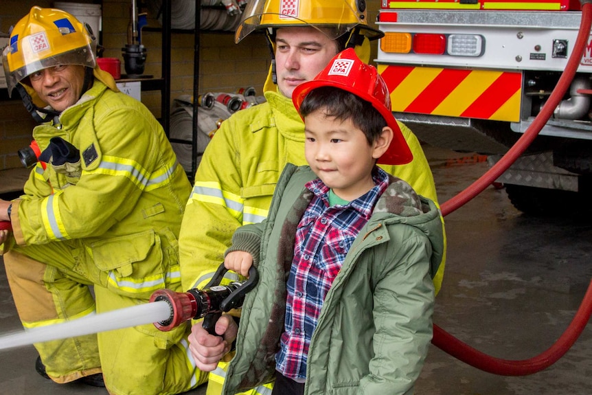A young boy wearing a fire helmet uses a hose attached to a fire truck with the help of uniformed fire fighters.
