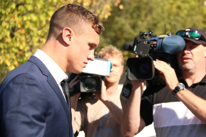 Jack Wighton, in a suit, walks with his head down while cameramen record him.