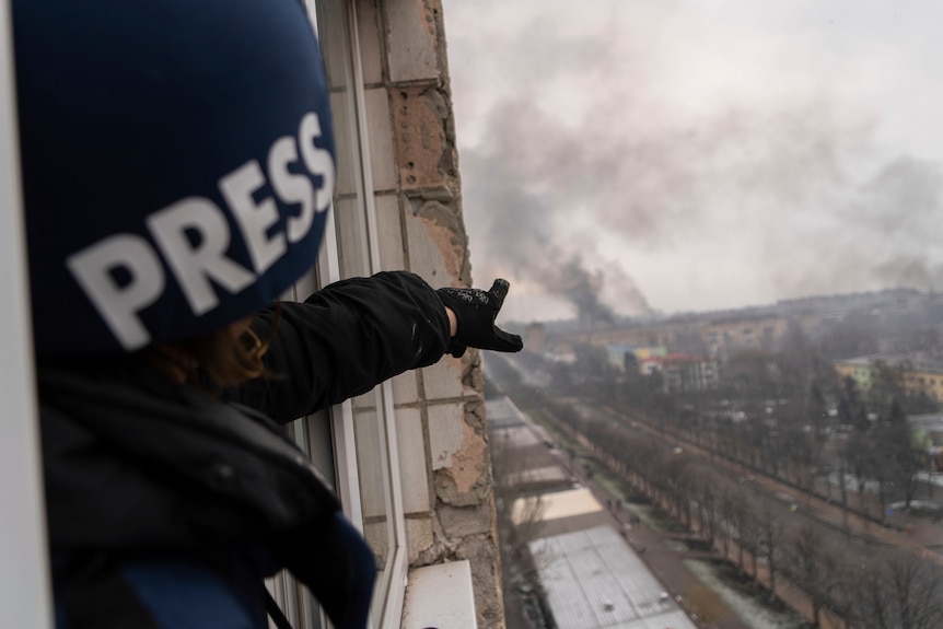 A journalist points at smoke rising over a city.