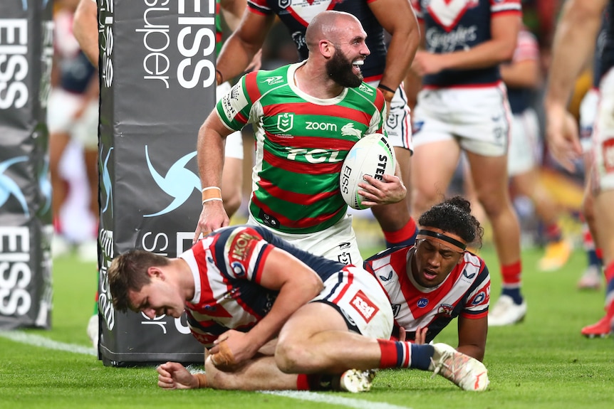 A South Sydney NRL player holds the ball while kneeling after scoring a try against the Roosters.