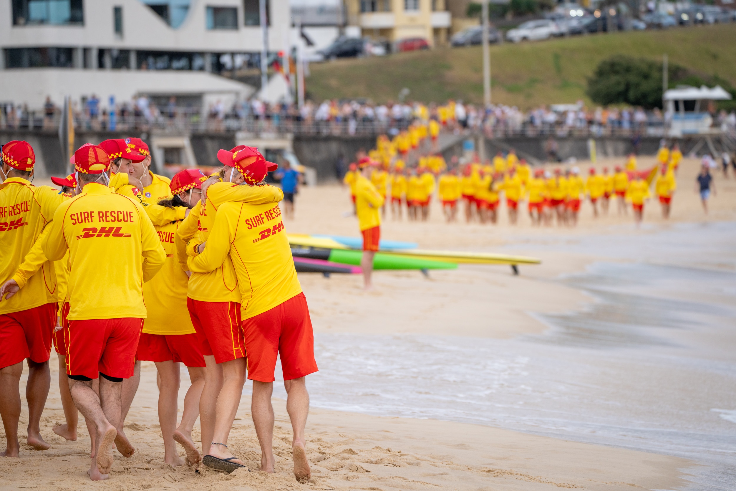 Vários salva-vidas se abraçam na praia. Ao longe, mais salva-vidas descem as escadas, algumas pranchas de surf colocadas no chão