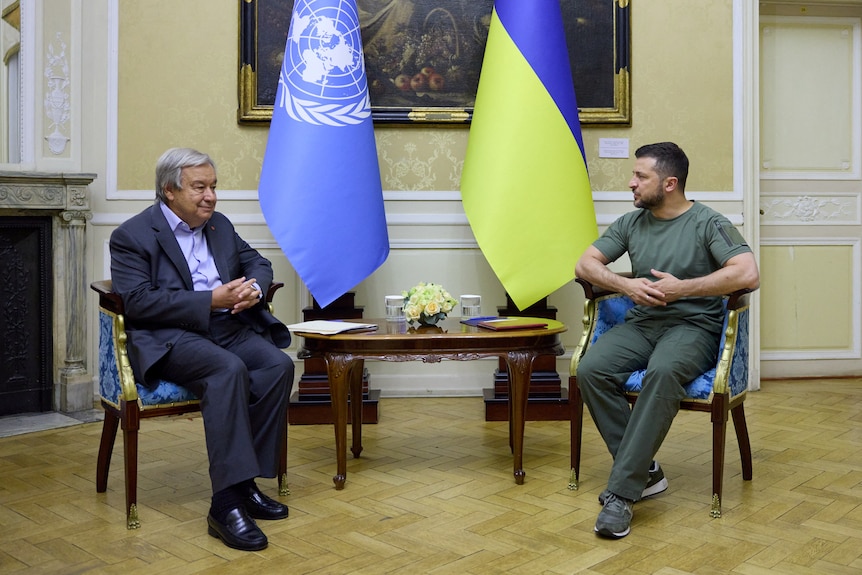 Two men sit opposite small table with UN and Ukrainian flags behind them.