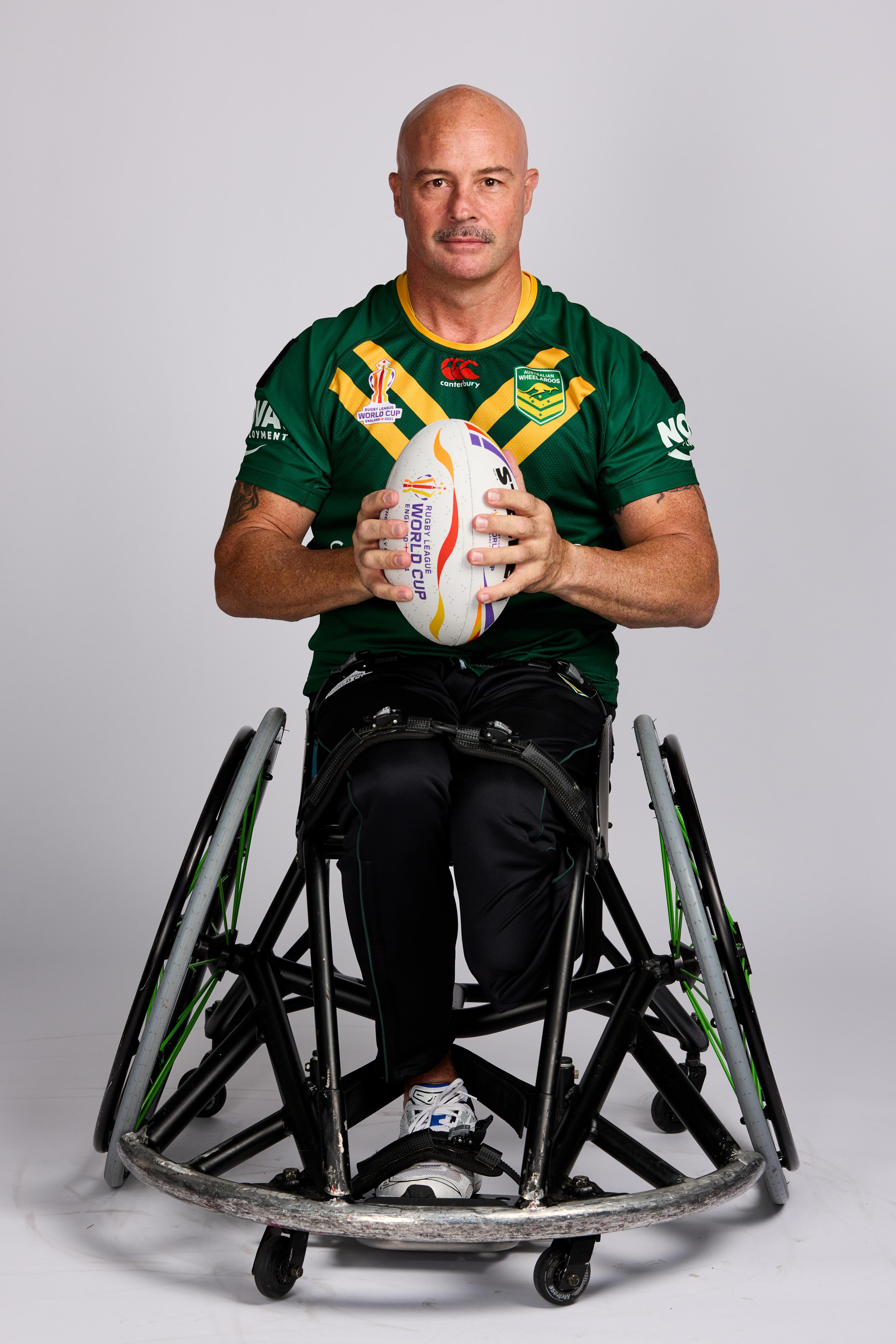 A bald man in a green shirt sits in a sports wheelchair and holds a rugby ball