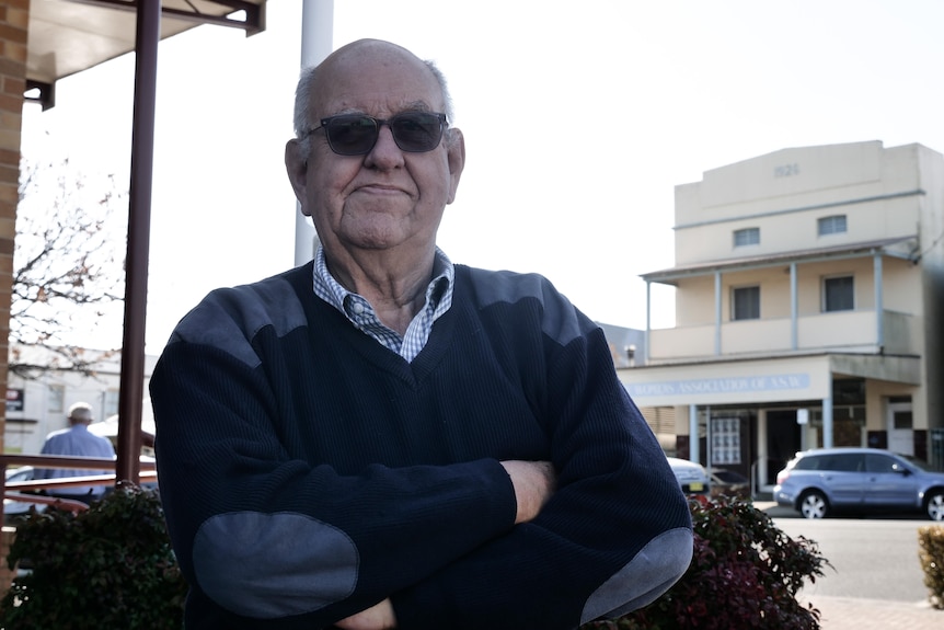 A serious bald, elderly man wears sunglasses, stands with his arms crossed, a cream Victorian building, cars, a man behind.