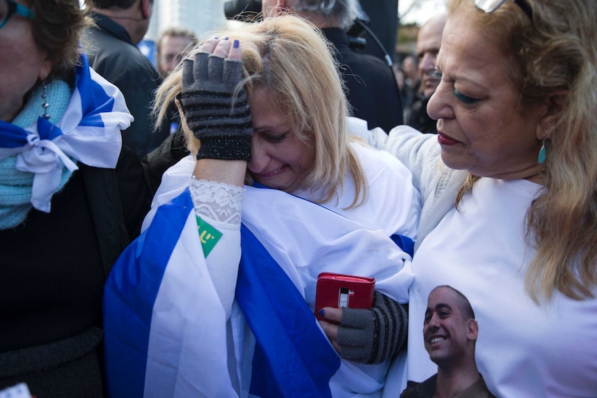 A woman wrapped in the Israeli flag holds her hand to her face as she cries.