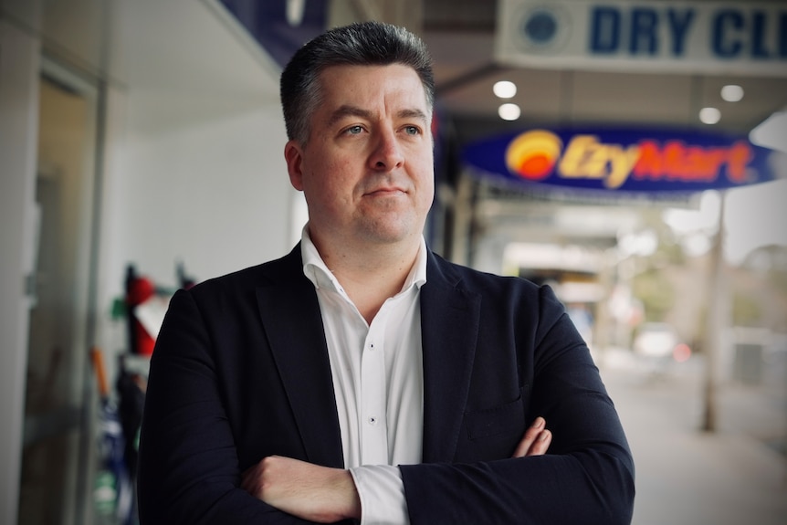 Luke Achterstraat stands with his arms crossed, with retail signage behind him.