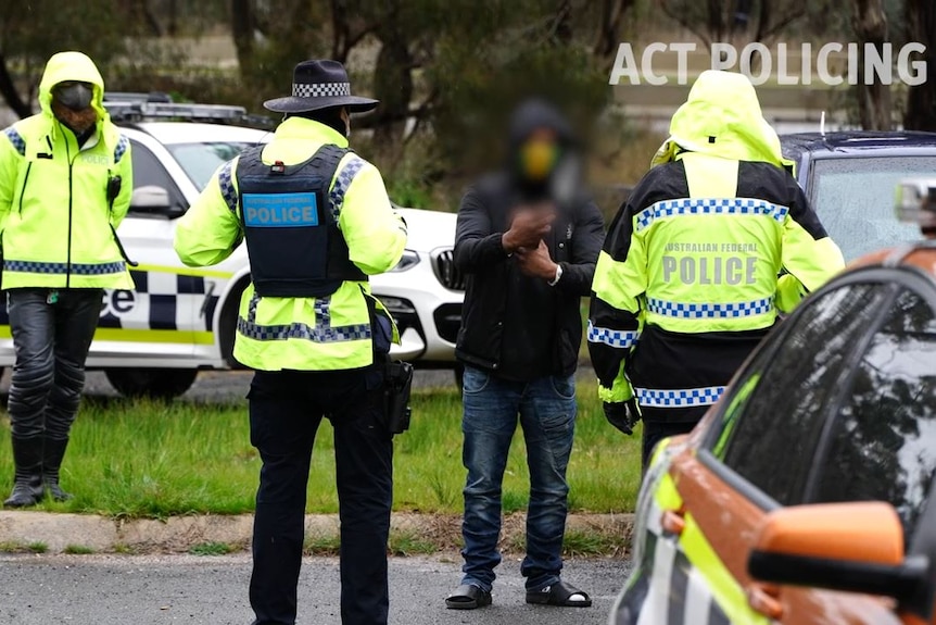 Three police officers in fluro, wet-weather gear talk to a man whose face is blurred.