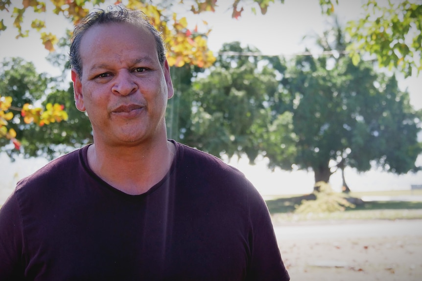 A man in a maroon shirt stands in front of trees and the ocean in the background