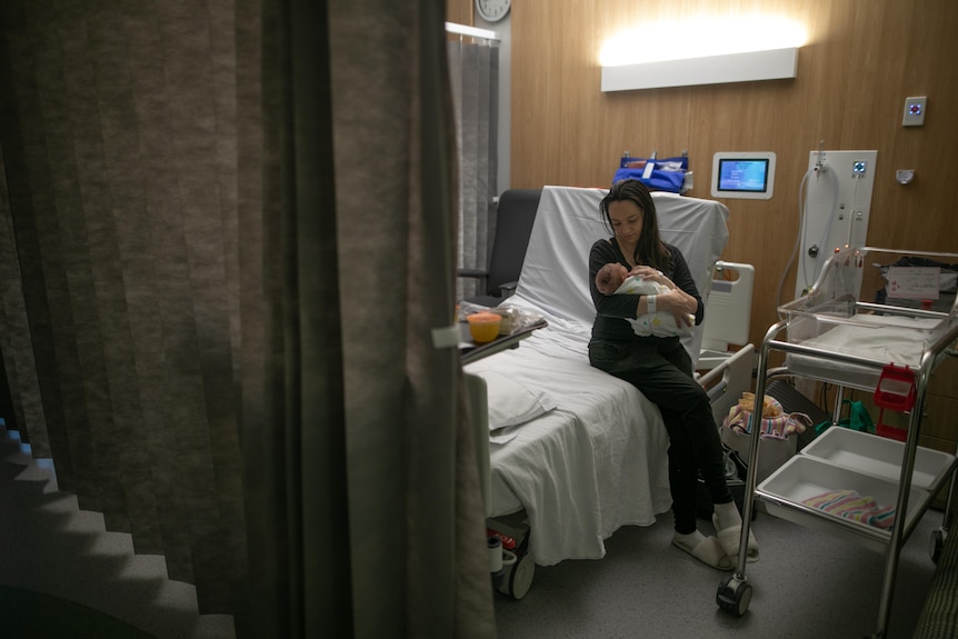 Taryn sits alone on a hospital bed, holding her newborn baby Darci in her arms.