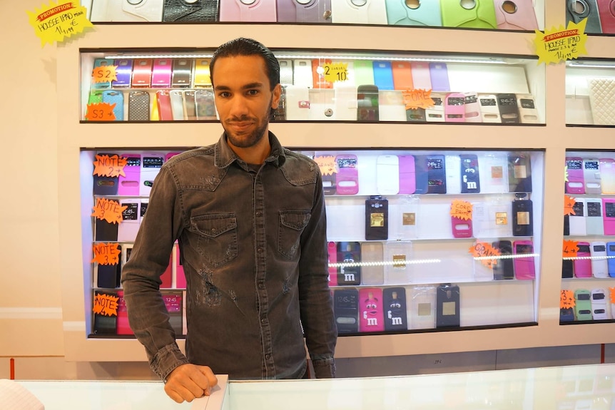 Issam stands in front of rows of mobile phone accessories in the store.