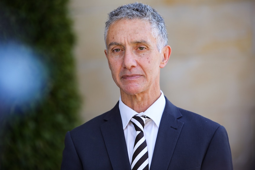 A slim man with short, grey hair, wearing a dark suit and speaking outside.