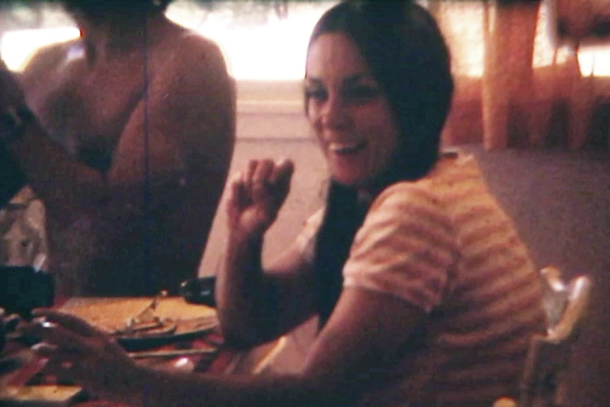 A grainy home video still showing a woman with brown pigtails sitting at a dining table and laughing with others.