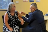 A woman in a dark dress sits on a chair while a man gives her an injection.