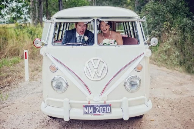 A bride and groom sit in the front of a 15 window Kombi van.