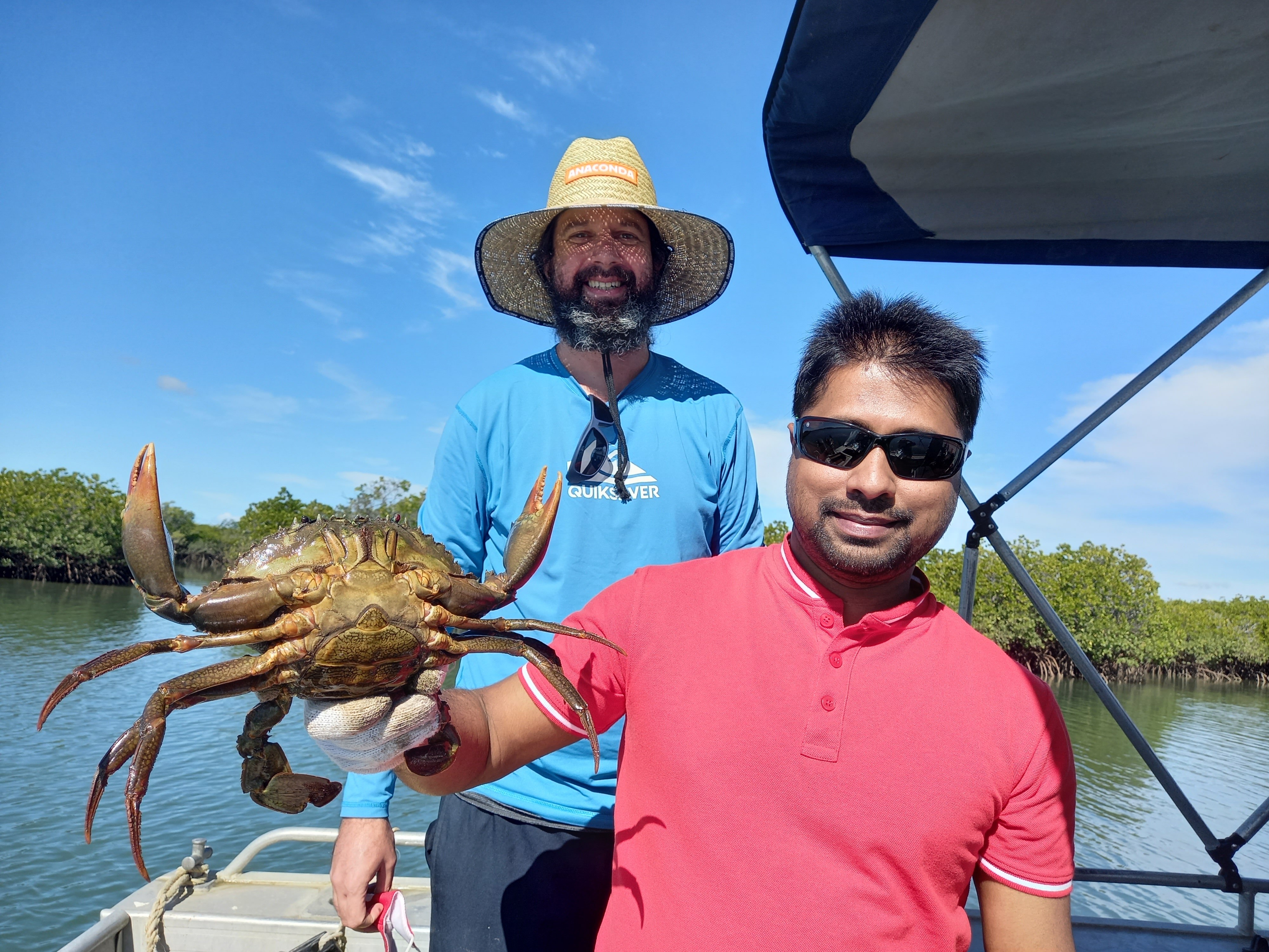 Two men standing on a boat, one wearing sunglasses, the other a hat, holding a giant mudcrab.