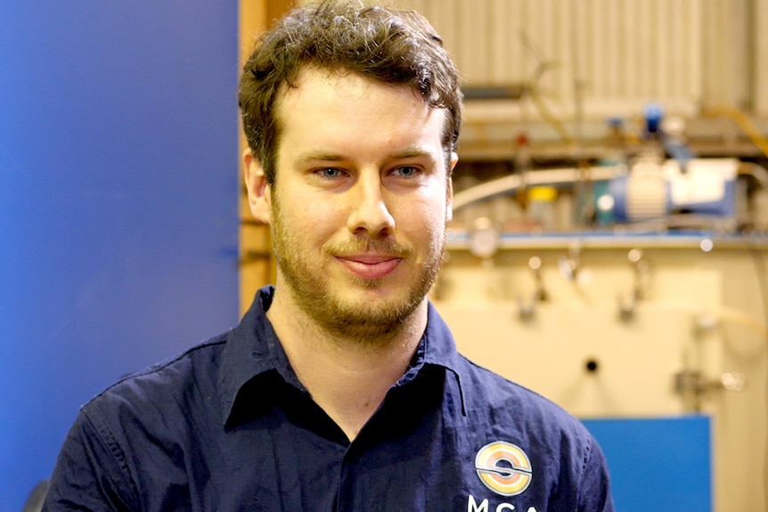 A man in a blue shirt labelled 'MGA' talks inside a lab.