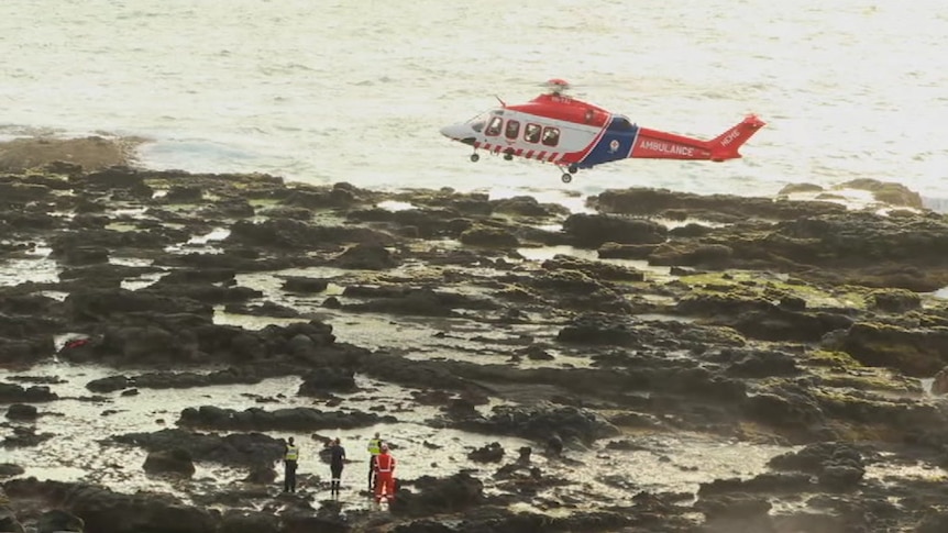 A helicopter hovers above rocks on the coast.