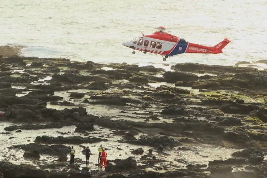 A helicopter hovers above rocks on the coast.