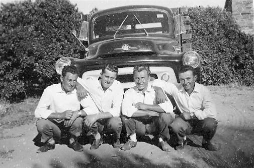 A black and white photo from the 1950s of four young men, all in white shirts and dark trousers, crouching in front of a car.
