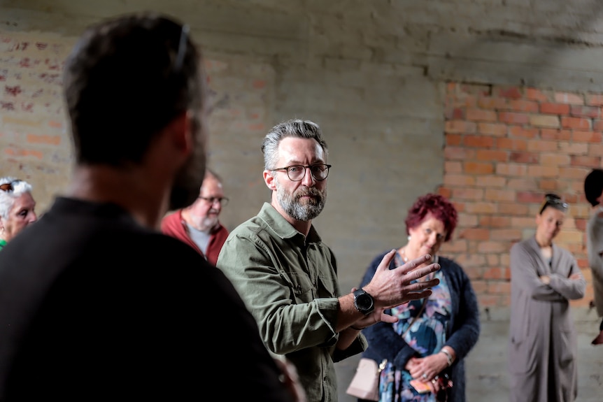 Man with glasses and beard in centre of a group of watching people inside a dilapidated factory gestures with hands