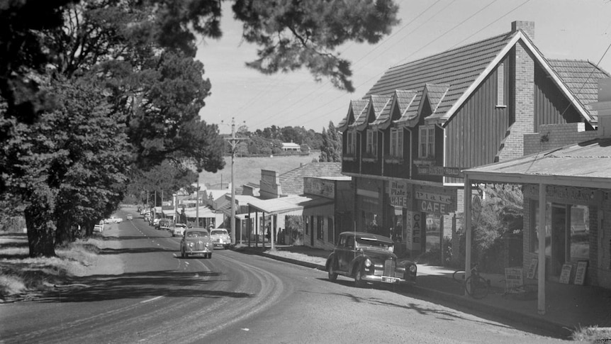 A black and white photograph of a country town road with trees left and buildings right