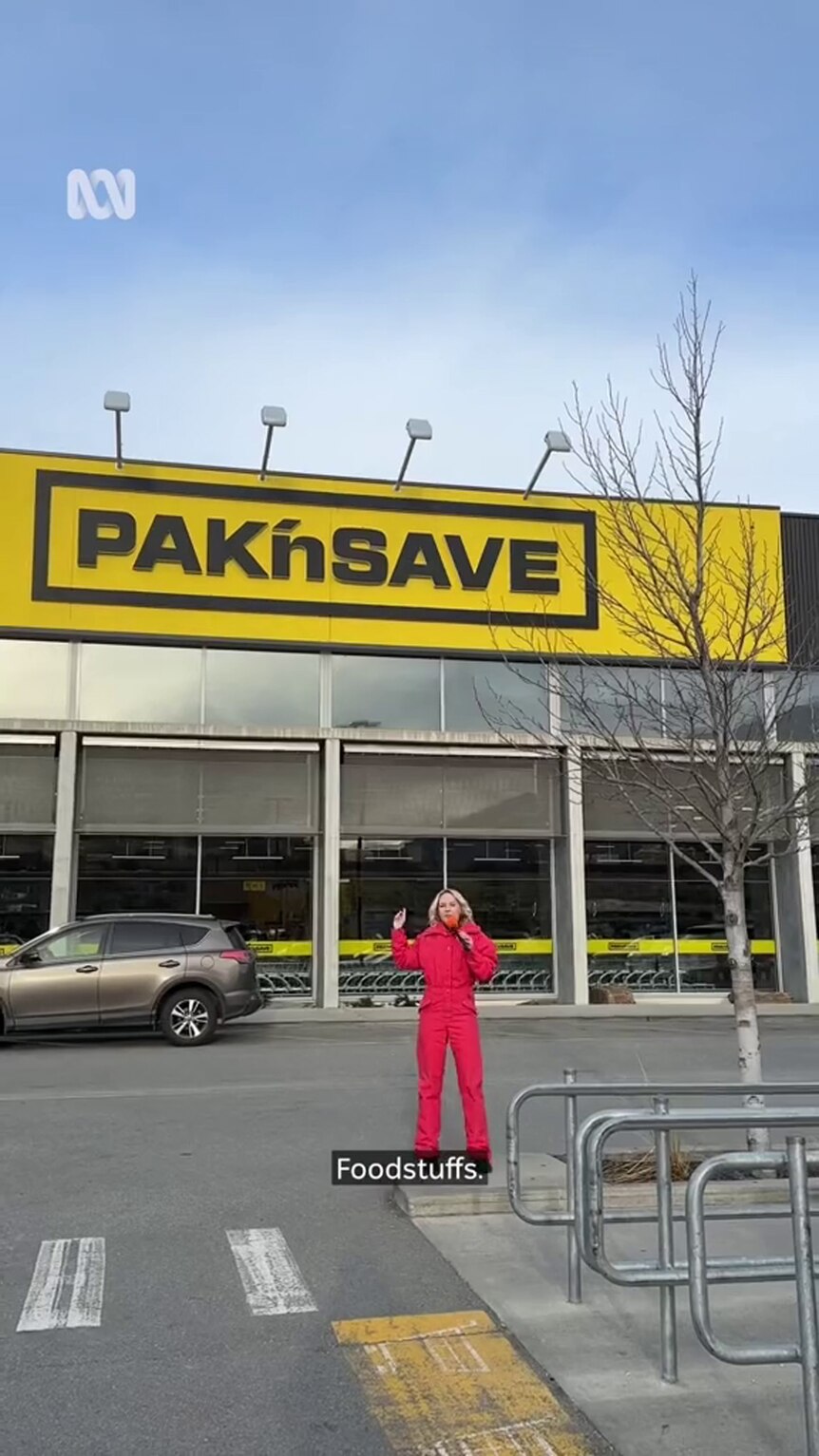 A young woman light-tone skin in a red snowsuit stands in a car park in front of a PAK'nSAVE 