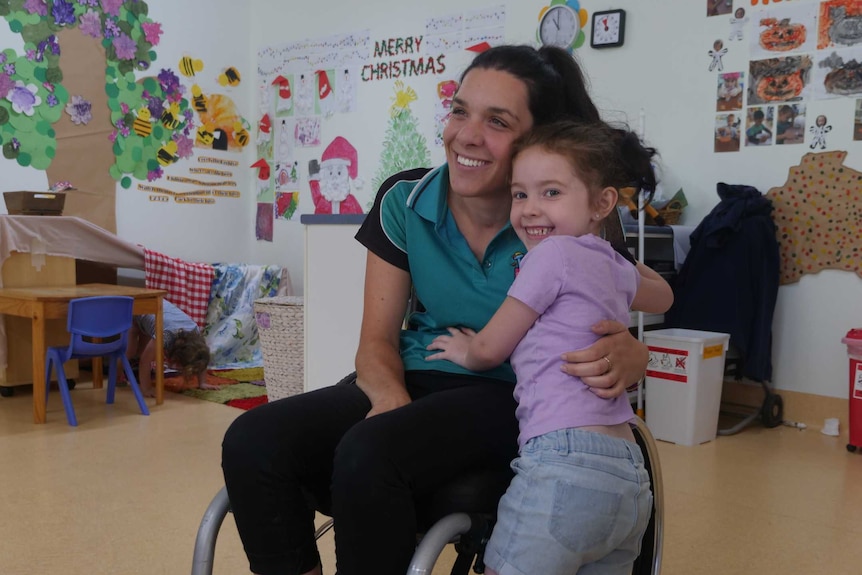 A lady in a wheelchair hugs a young girl in a preschool room.