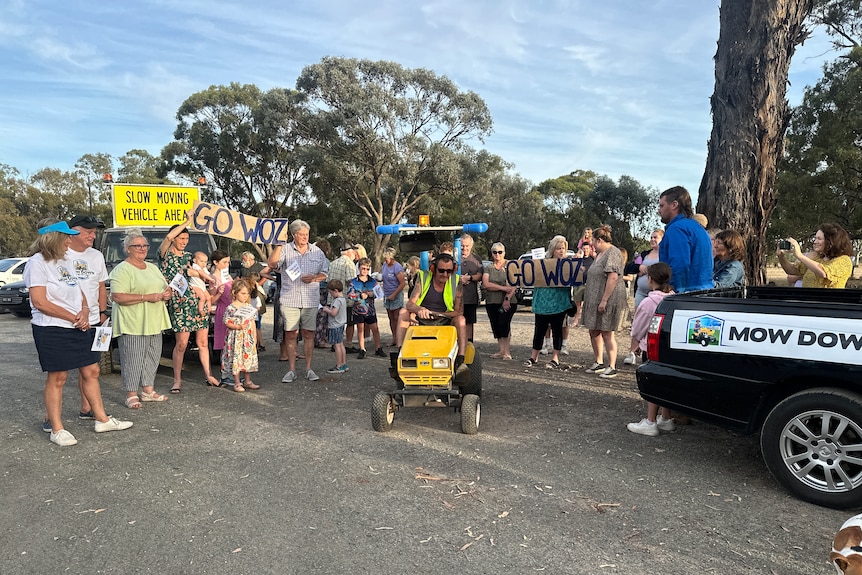 A crowd of people watch on as a man in high-vis steers a ride-on mower onto a road.