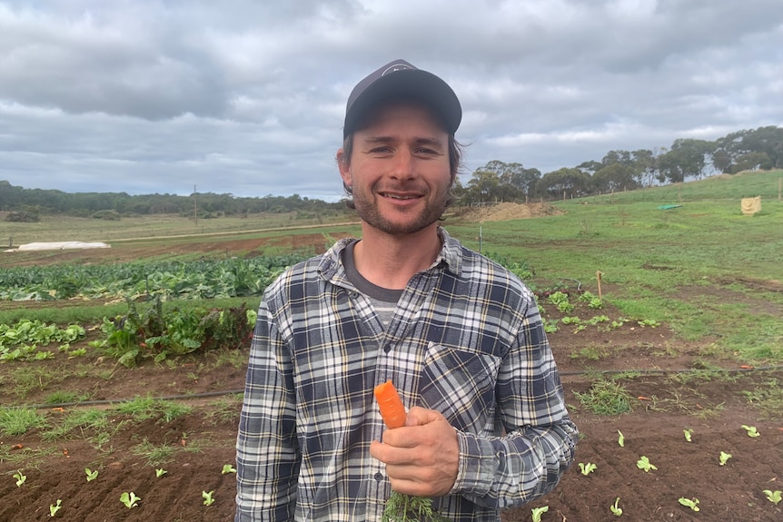 A happy man standing in front of a vegetable farm eating a carrot