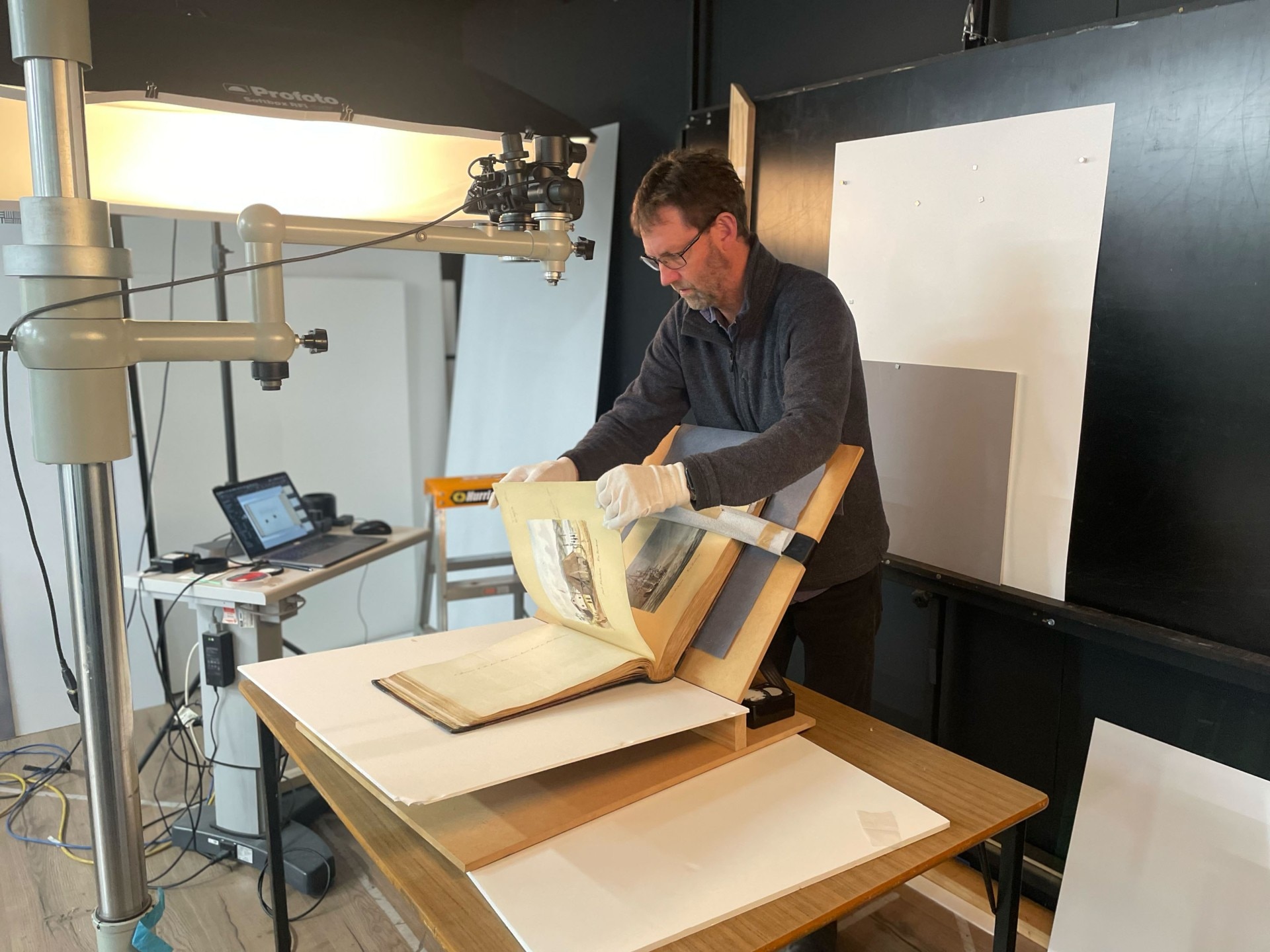 A man wearing a blue top and gloved hands turns a page of an old album, with a camera overhead. 