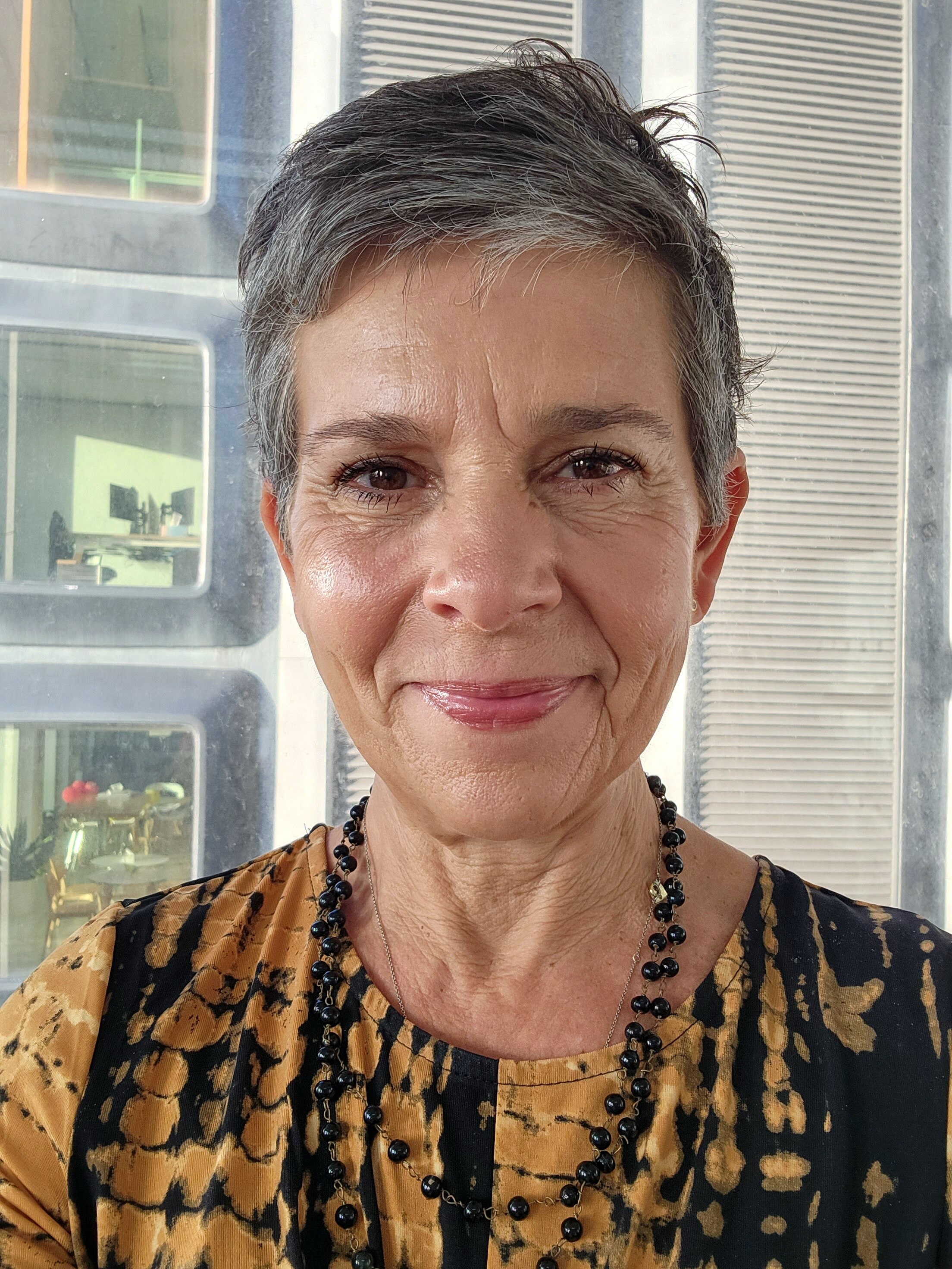 A smiling woman with short graying hair wearing an orange and black top and brown bead necklace.