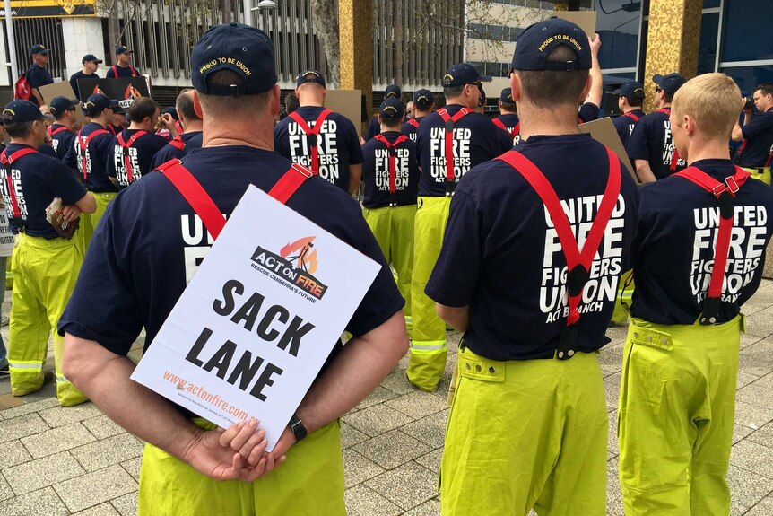 A group of ACT firefighters, pictured from the back, at a rally outside the ACT Legislative Assembly. One sign reads "SACK LANE"