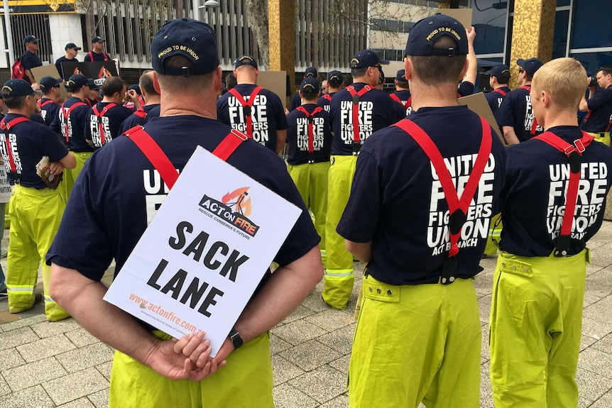 A group of ACT firefighters, pictured from the back, at a rally outside the ACT Legislative Assembly. One sign reads "SACK LANE"