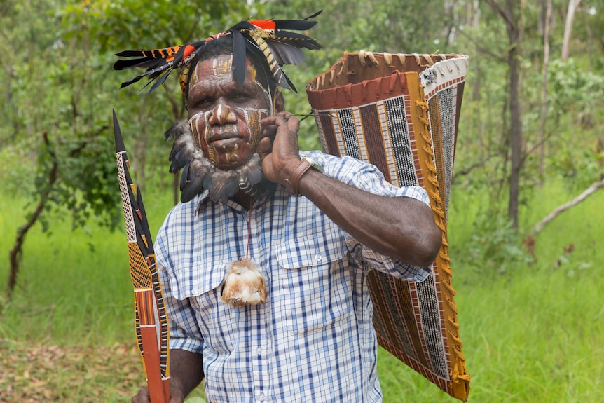 Pedro Wonnaeamirri stands in bushland holding art work and wearing face paint.