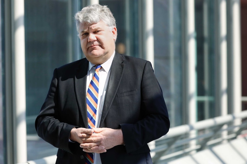 A grey-haird man in a black suit and blue and orange tie stares into the camera with his hands clasped in front of him.