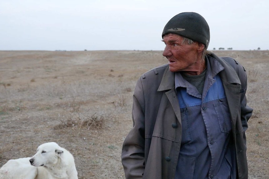 Boris Dainix walks with a stick across barren land accompanied by a dog